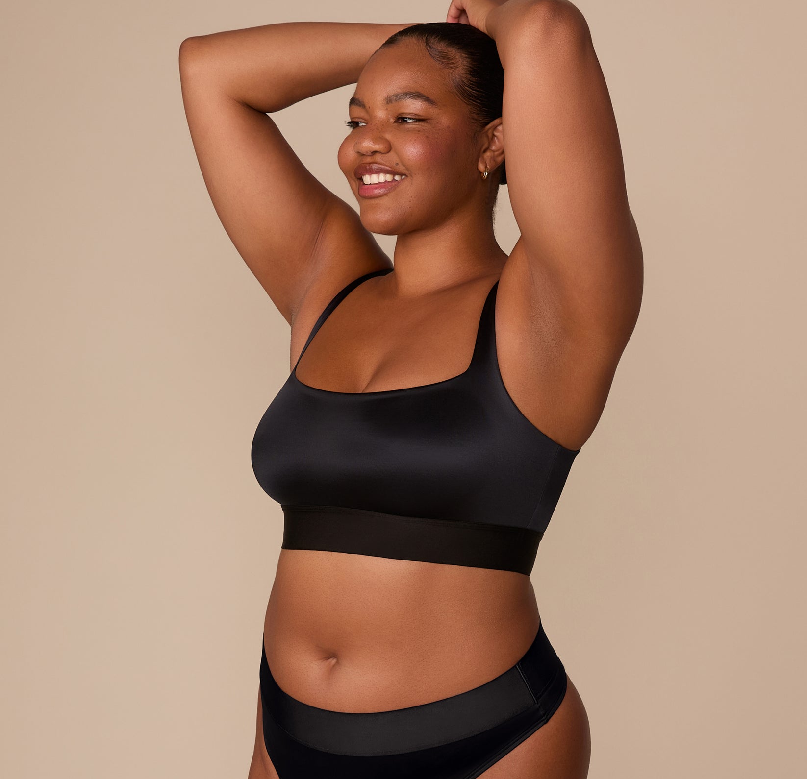 Zarria smiling with arms raised, wearing the Stretch Satin Square Neck Bralette in Black. The satin finish and extended underband are clearly visible.