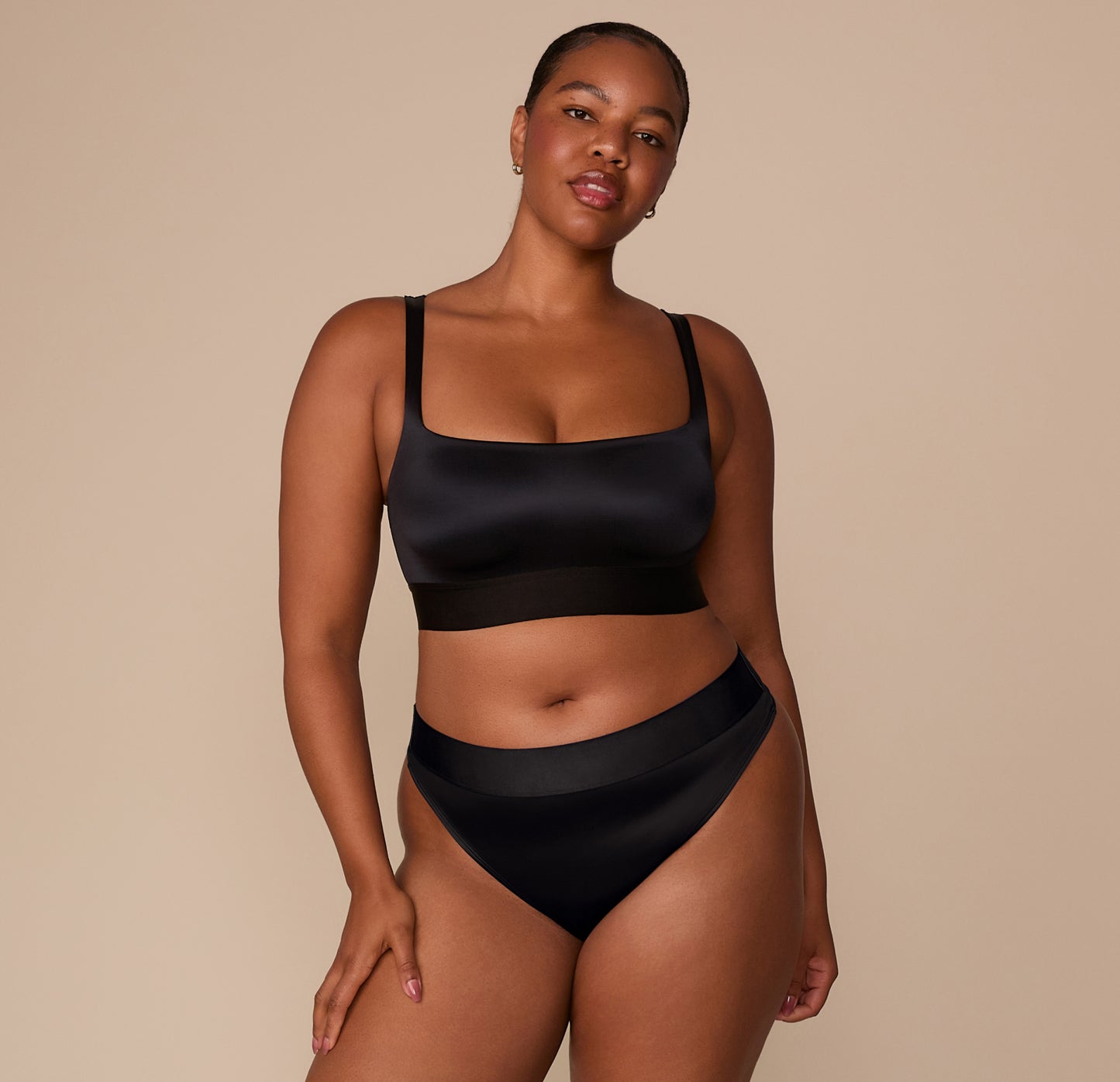 Full front view of Zarria in the Stretch Satin Square Neck Bralette and coordinating Stretch Satin Highwaist Thong in Black, with a confident, relaxed pose.
