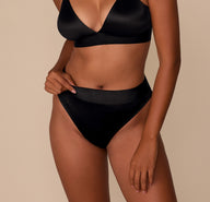 Victoria wearing the Stretch Satin Highwaist Thong in Black with the matching Stretch Satin Triangle Bralette, standing in a relaxed pose. The mid-rise thong fit is visible from the front. sizing: Victoria wears size Medium