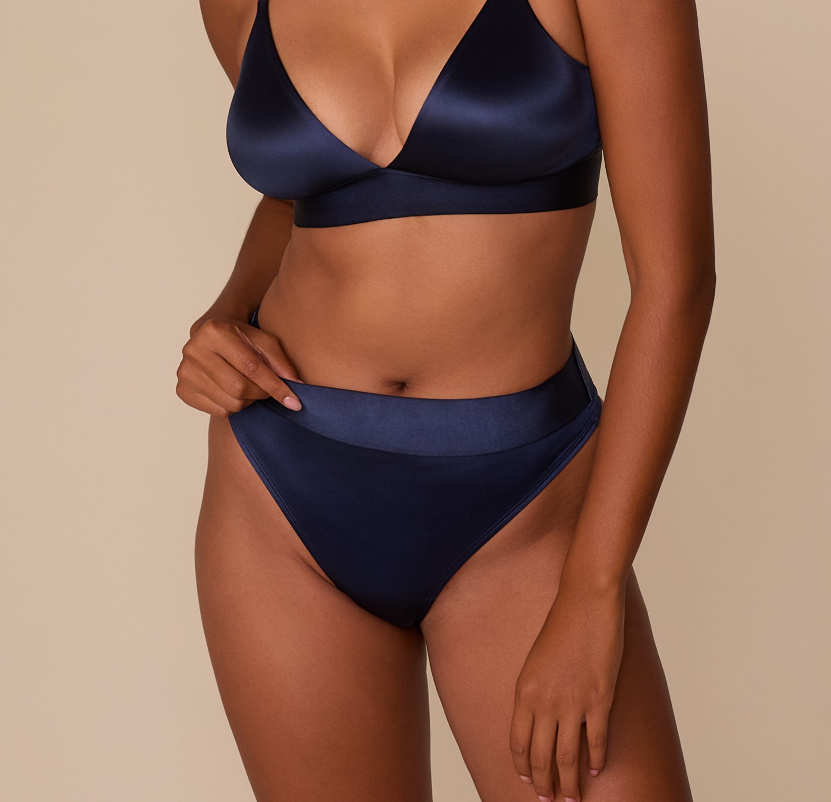 Front view of Victoria wearing the Stretch Satin Highwaist Thong in Navy, lightly pinching the waistband to show fit and flexibility. sizing: Victoria wears size Medium