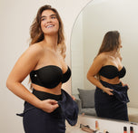 Summerlyn stands in front of a mirror in a well-lit room, wearing the Base Strapless Bra and No-Show Highwaist Brief in Black. She is smiling and adjusting her navy dress, with makeup items visible on the counter, getting ready for an event.