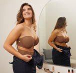 Summerlyn stands in front of a mirror, wearing the Base Strapless Bra and No-Show Highwaist Brief in Brown. She is smiling while adjusting her navy dress, capturing a moment of getting ready for an event.