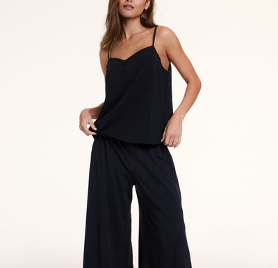 In a studio with a neutral backdrop, Sunday wears the Sunday Pima Cami Pant Set in Black, featuring a lightweight camisole with thin adjustable straps and a relaxed fit, paired with wide-leg pants made from soft, breathable fabric. She gently pulls the hem of the top to highlight the easy, comfortable fit.