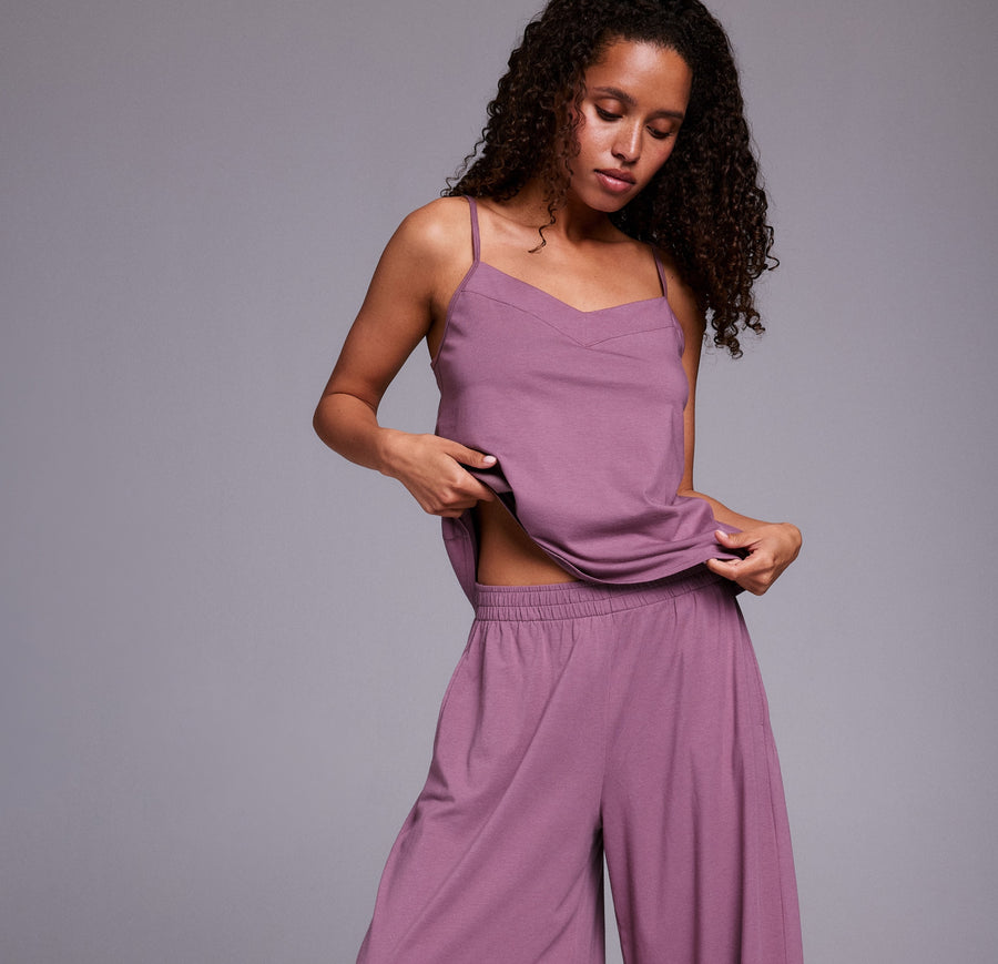 In a studio with a neutral backdrop, Sunday wears the Sunday Pima Cami Pant Set in Mirage, featuring a lightweight camisole with thin adjustable straps and a relaxed fit, paired with wide-leg pants made from soft, breathable fabric. She lifts the hem of the top to reveal the comfortable elastic waistband.