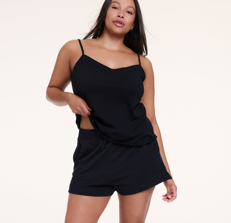 In a studio with a neutral backdrop, Sunday wears the Sunday Pima Cami Short Set in Black, featuring a lightweight camisole with thin adjustable straps and a relaxed fit, paired with soft, breathable shorts with an elastic waistband. She lifts the hem of the top slightly to show the comfortable fit.