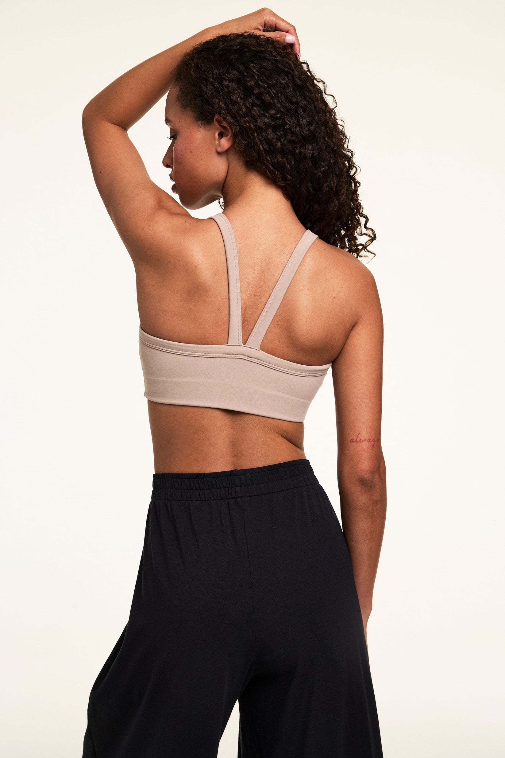 Back view on a light studio backdrop of Courtney wearing the Whipped Knit V-Neck Bralette in Mushroom with black wide-leg lounge pants. The bralette features wide straps, a V-back design, and a smooth longline band.