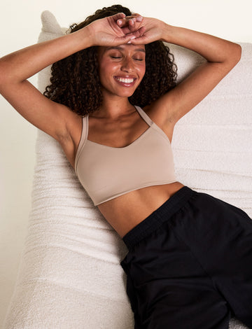 In a relaxed indoor setting, Courtney reclines on a cream lounge chair wearing the Whipped Knit V-Neck Bralette in Mushroom with black lounge pants. The bralette features a V-neck silhouette and soft knit construction designed for everyday comfort. sizing: Courtney wears size Small