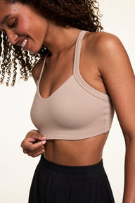 Side close-up on a light studio backdrop of Courtney wearing the Whipped Knit V-Neck Bralette in Mushroom with black lounge pants. Courtney gently holds the hem of the bralette, showing the smooth knit fabric and longline band.