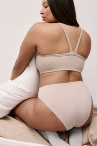 In a softly lit bedroom, Alana wears the Whipped Knit V-Neck Bralette in Mushroom with matching underwear, shown from the back while seated and holding a pillow. The bralette features a racerback-style strap detail and a smooth, wireless band for comfortable support.