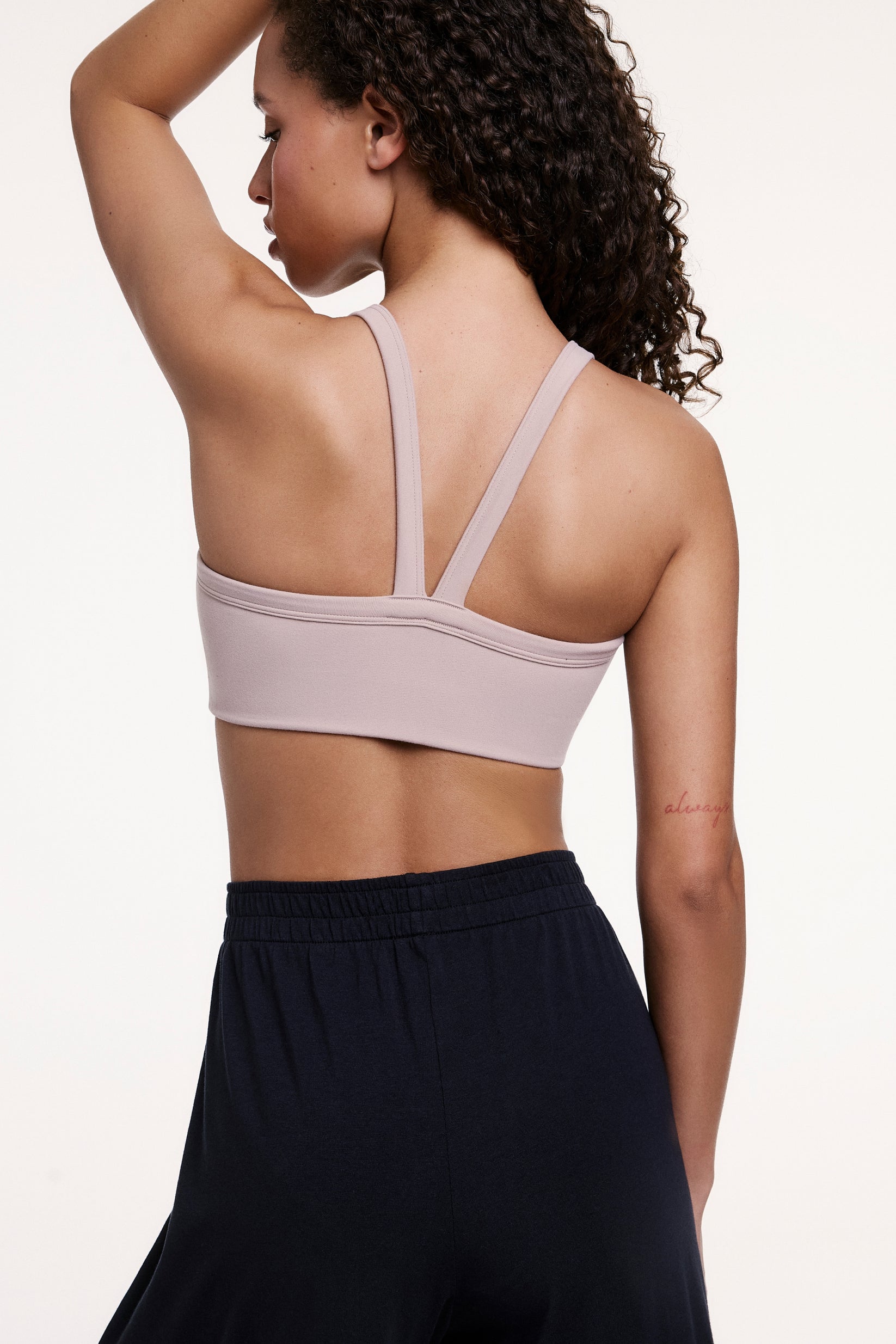 In a studio setting, Courtney wears the Whipped Knit V-Neck Bralette in Mushroom, shown from the back with one arm raised. The bralette features a racerback-style strap detail and a smooth, wireless band for comfortable support.