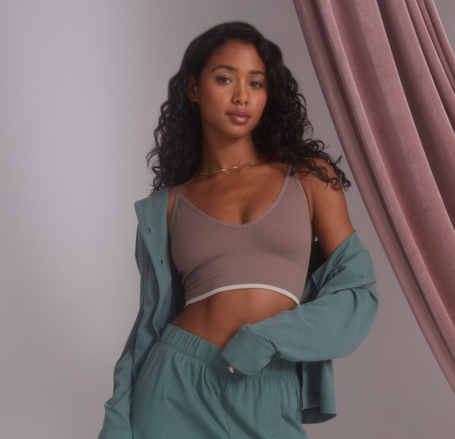 Ally is standing with a soft gaze, wearing the Lounge Rib Triangle Bralette in Oyster, layered with an open matching Perfect Pima Sleep set in Rainforest green. The bralette features ribbed detailing and a subtle contrast trim along the bottom band. A flowing mauve curtain adds texture in the background, creating a relaxed and cozy atmosphere.
