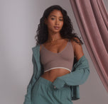 Ally is standing with a soft gaze, wearing the Lounge Rib Triangle Bralette in Oyster, layered with an open matching Perfect Pima Sleep set in Rainforest green. The bralette features ribbed detailing and a subtle contrast trim along the bottom band. A flowing mauve curtain adds texture in the background, creating a relaxed and cozy atmosphere.