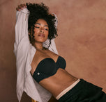 Model sitting in a chair with her arms raised and crossed behind her head. She's wearing the Base Strapless Bra in Black with an open button down beige shirt and gold chain necklace.