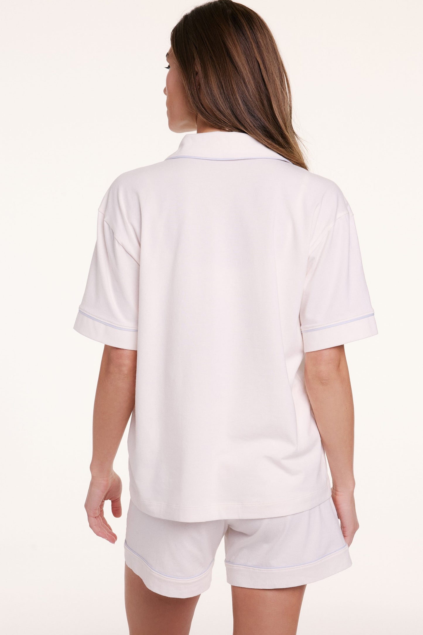 On a neutral backdrop, Gianna faces away wearing the Perfect Pima Pajama Shirt in Ivory with matching shorts, showing the relaxed fit and clean back silhouette with contrast piping along the sleeves.