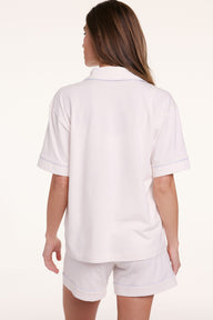 On a neutral backdrop, Gianna faces away wearing the Perfect Pima Pajama Shirt in Ivory with matching shorts, showing the relaxed fit and clean back silhouette with contrast piping along the sleeves.