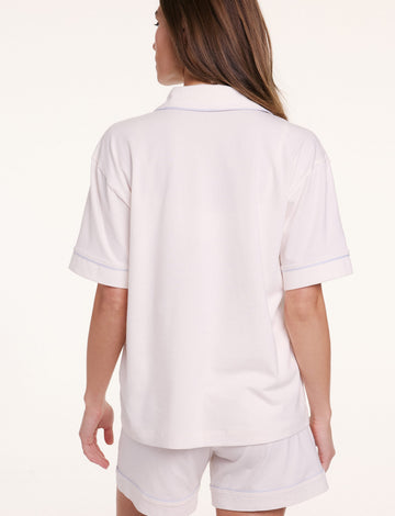 On a neutral backdrop, Gianna faces away wearing the Perfect Pima Pajama Shirt in Ivory with matching shorts, showing the relaxed fit and clean back silhouette with contrast piping along the sleeves.