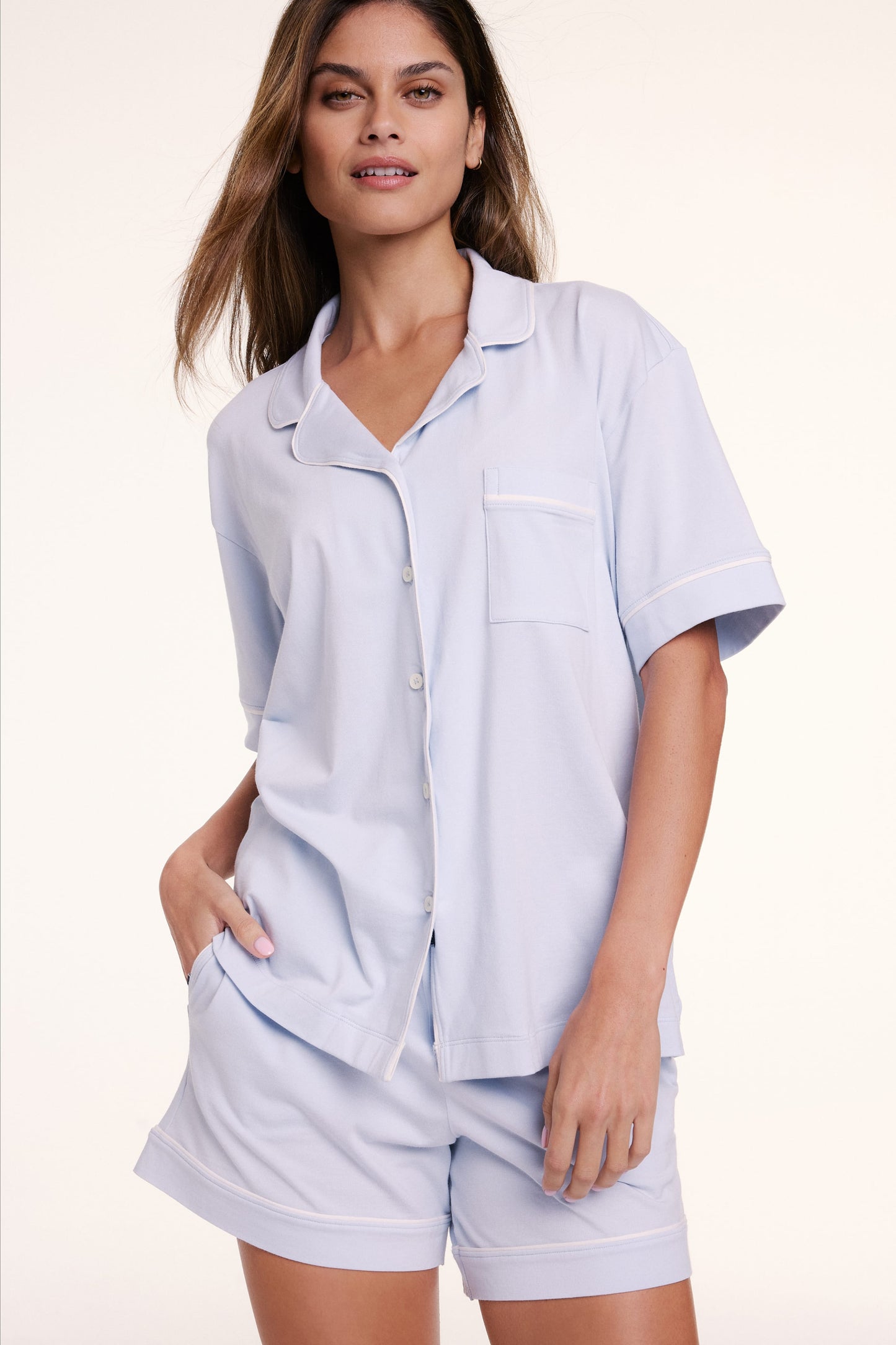 In a studio setting, Linni wears the Perfect Pima Pajama Short Sleeve Shirt in Soft Blue with matching shorts, featuring a button-front design, chest pocket, and soft, breathable fabric.
