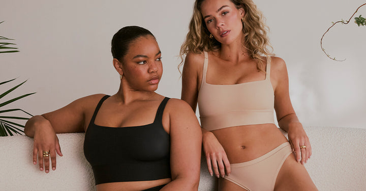 Zarria and Freya relax on a neutral-toned couch, wearing the Harper Wilde Bliss Square Neck Bralette and matching briefs. Zarria wears the set in Black, highlighting the sleek longline silhouette and smoothing fit. Freya wears the set in Beige, showcasing the soft, barely-there comfort of the fabric. Their effortless, joyful expressions and cozy setting emphasize the everyday ease and versatility of this new style.