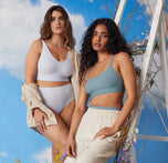 Two models standing against a blue sky backdrop. Summerlyn is standing wearing the Bliss Bralette and matching Bliss Highwaist Brief in Tidal. Naisha is leaning against a white ladder adorned in flowers wearing the Bliss Scoop in Mist with off white linen pants.