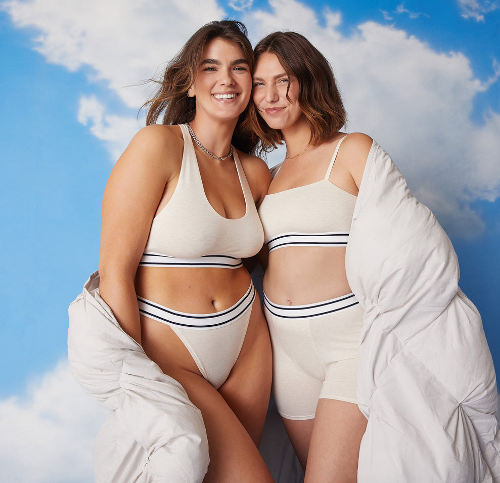 Two models wrapped in a puffy white-gray quilt against a blue sky cloudy backdrop. Summerlyn is wearing the Cloud Cotton Logo Racerback Bralette and matching Cloud Cotton Logo French Cut Bikini in Heather Oatmeal. Carli is wearing the Cloud Cotton Logo Bandeau and matching Cloud Cotton Logo Boxer Brief in Heather Oatmeal.