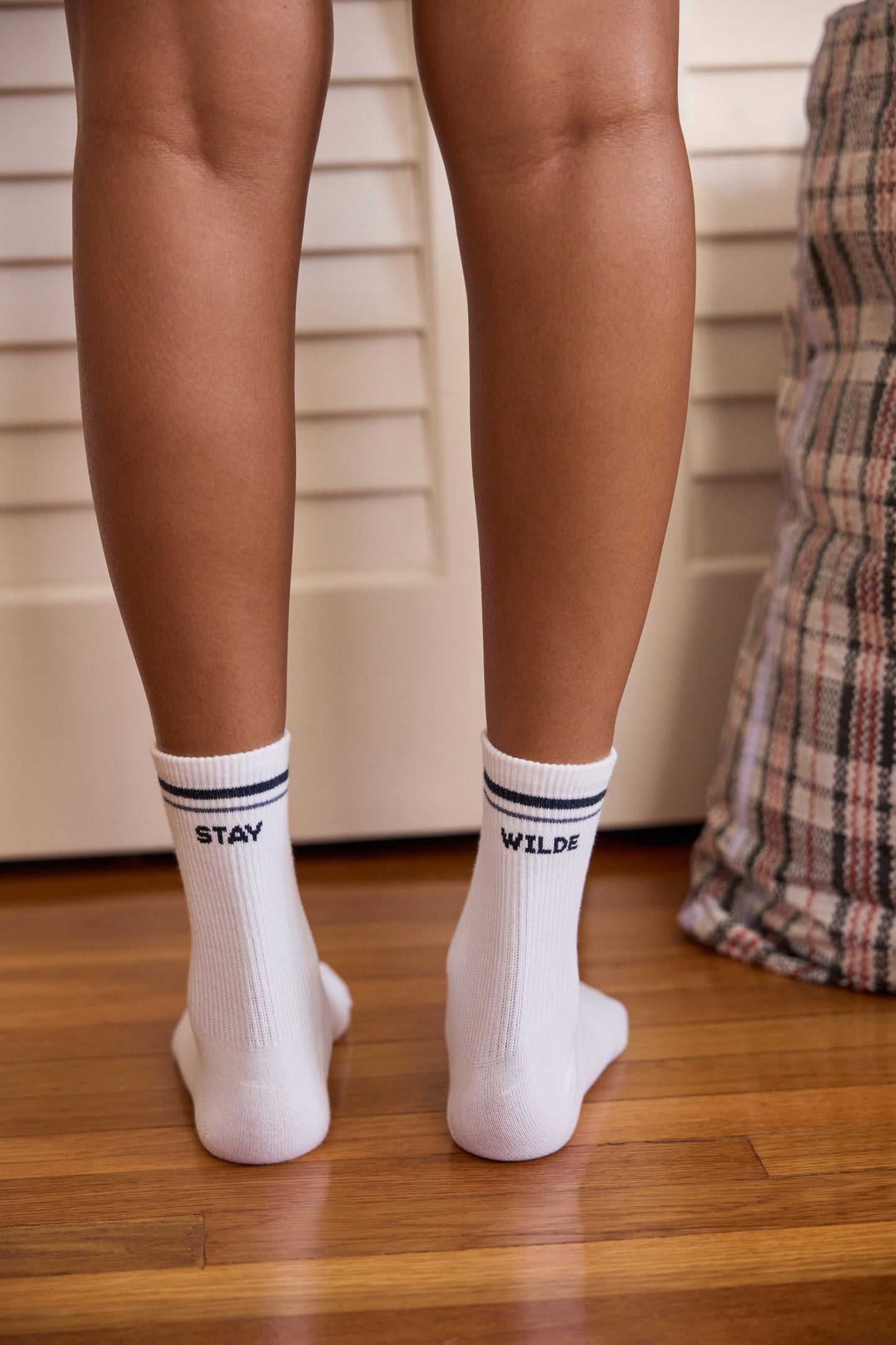 Back view of white crew socks with navy stripe trim, reading 'STAY WILDE' across both socks, worn on a model standing on a wood floor.