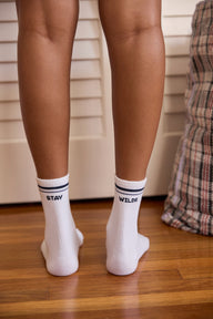 Back view of white crew socks with navy stripe trim, reading 'STAY WILDE' across both socks, worn on a model standing on a wood floor.