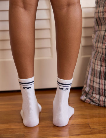 Back view of white crew socks with navy stripe trim, reading 'STAY WILDE' across both socks, worn on a model standing on a wood floor.
