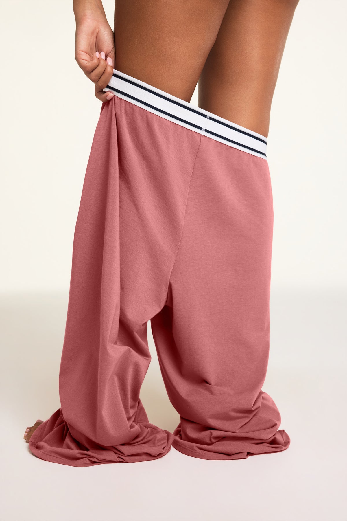 A model wearing dusty rose straight cotton pants with a contrasting white elastic waistband featuring thin navy stripes, shown from the waist down against a plain light background.