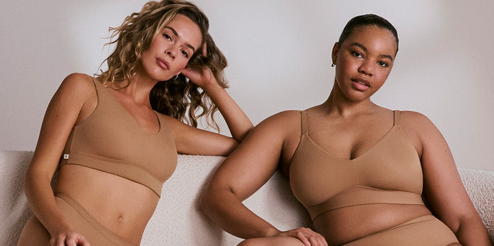 Two models of varying skin tones and size lounge on a cream colored sofa wearing the Bliss Bralette and Bliss Triangle Bralette with the matching Bliss Highwaist Brief in Tan. The bralette's soft underband and microfiber fabric is emphasized.