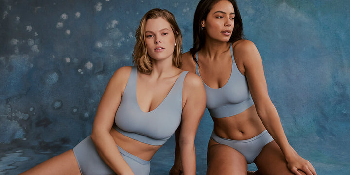 Two models pose against a rich blue backdrop. The model on the left wears the Bliss Bralette and Bliss Highwaist Brief in Glacier, while the model on the right wears the Bliss Racerback Bralette and Bliss Bikini in Glacier. Both styles feature a soft, seamless design in a cool, airy blue tone.