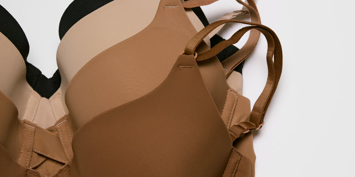 On a light studio background, several bras in neutral shades are layered together, showing smooth molded cups, soft microfiber fabric, and thin adjustable straps in tones of black, beige, tan, and brown.