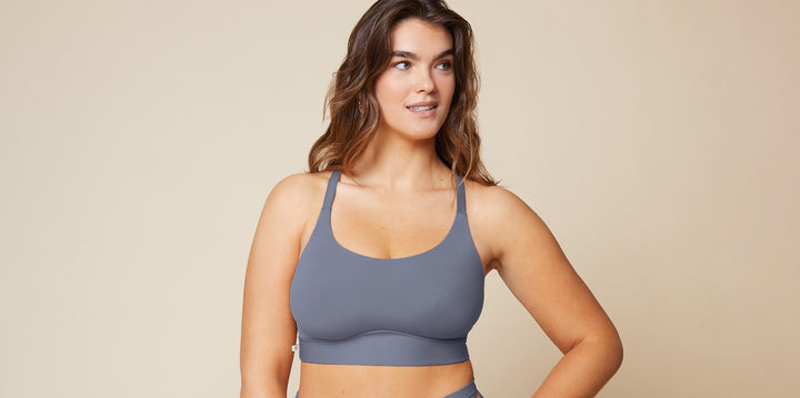 In a studio setting with a neutral beige background, a model wears a slate blue wireless scoop bralette with wide straps and a smooth band, standing with one hand on her hip.