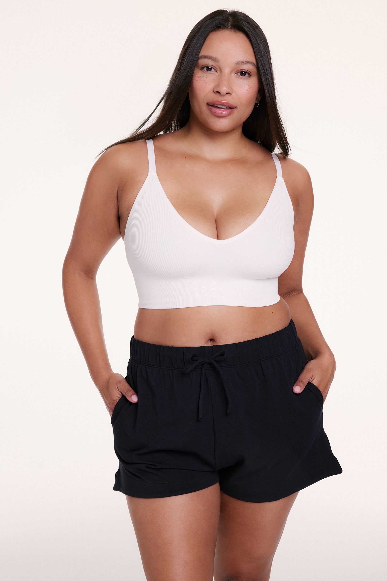 On a white studio backdrop, Alana wears the Sunday Pima Short in Black with a white bralette, featuring a relaxed fit, elastic waistband, and adjustable drawstring for a comfortable, easy feel. sizing: Alana wears size Large