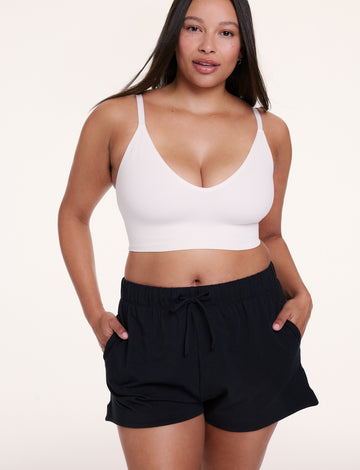 On a white studio backdrop, Alana wears the Sunday Pima Short in Black with a white bralette, featuring a relaxed fit, elastic waistband, and adjustable drawstring for a comfortable, easy feel. sizing: Alana wears size Large