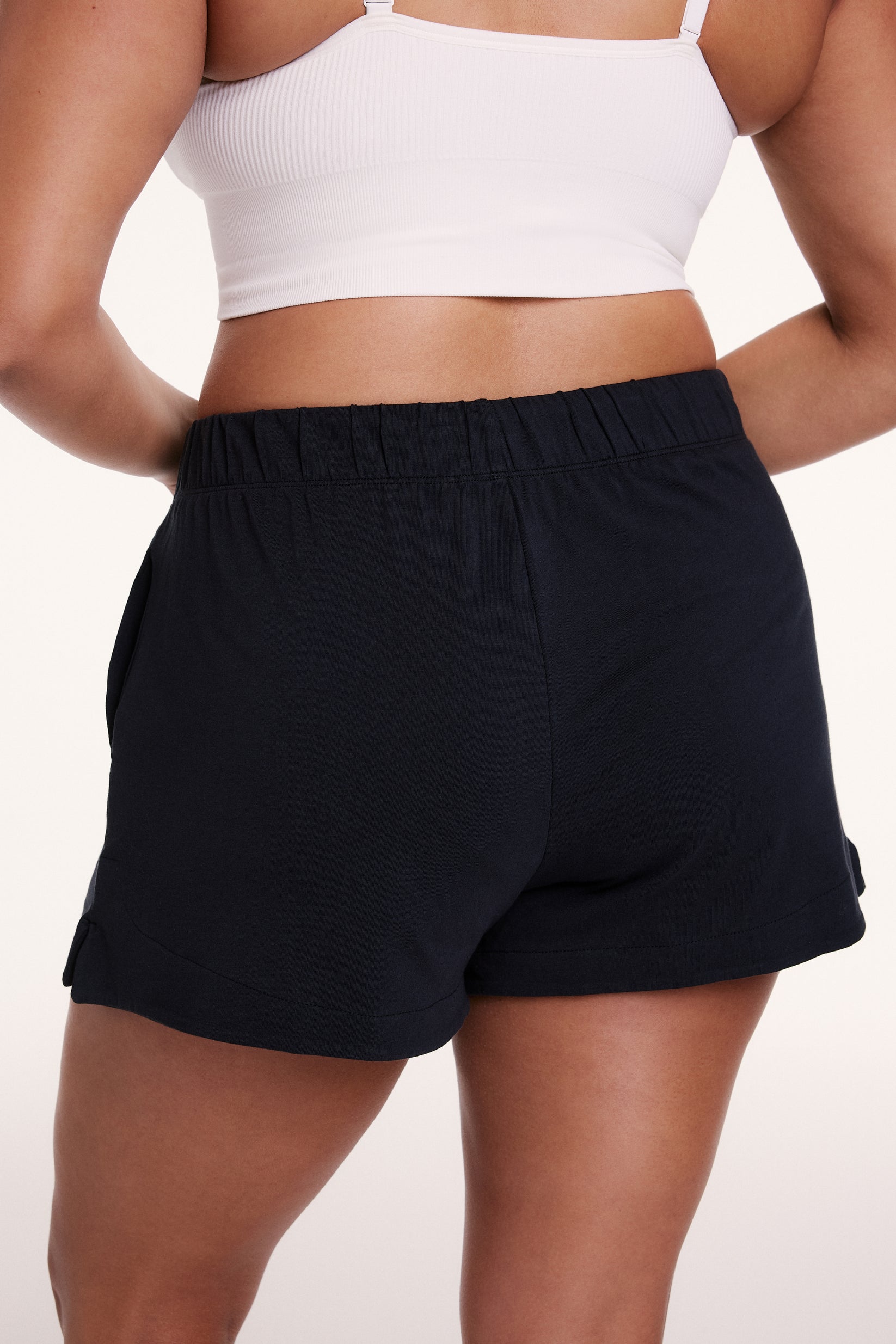 On a white studio backdrop, Alana wears the Sunday Pima Short in Black, showing the relaxed fit, elastic waistband, and smooth, lightweight fabric from the back.