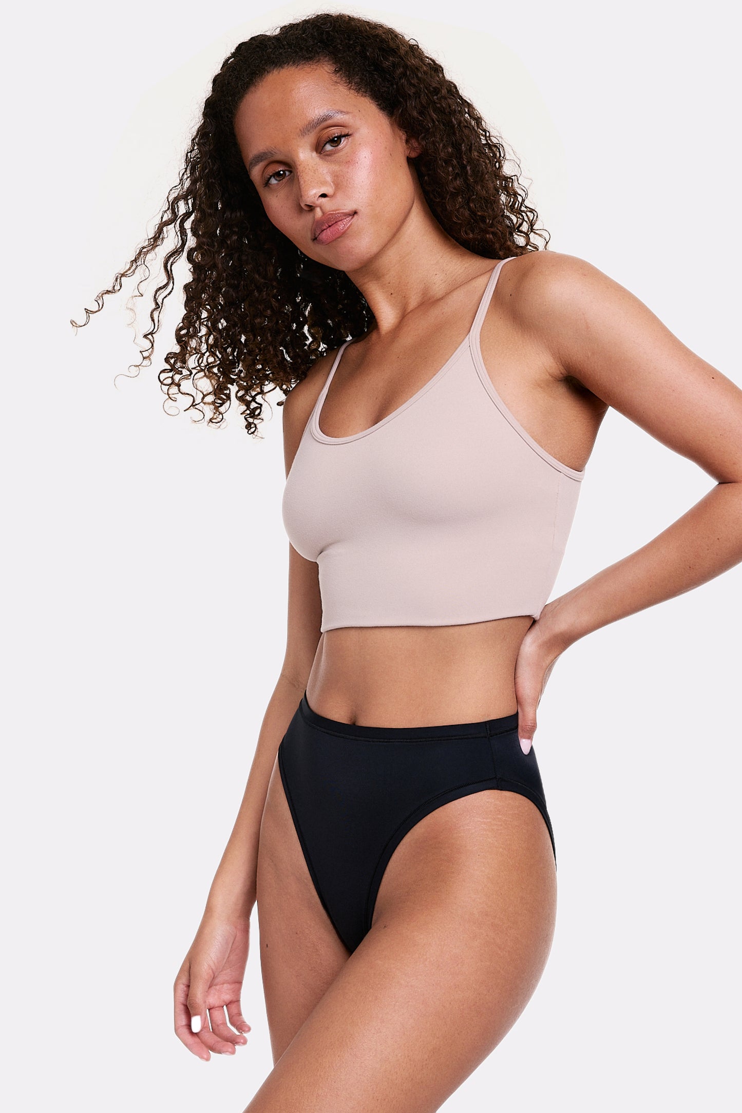 In a light gray studio, Courtney wears the Whipped Knit Scoop Bralette in Mushroom with black underwear, shown in a three-quarter side view highlighting the scoop neckline, thin adjustable straps, and soft, smoothing fabric.