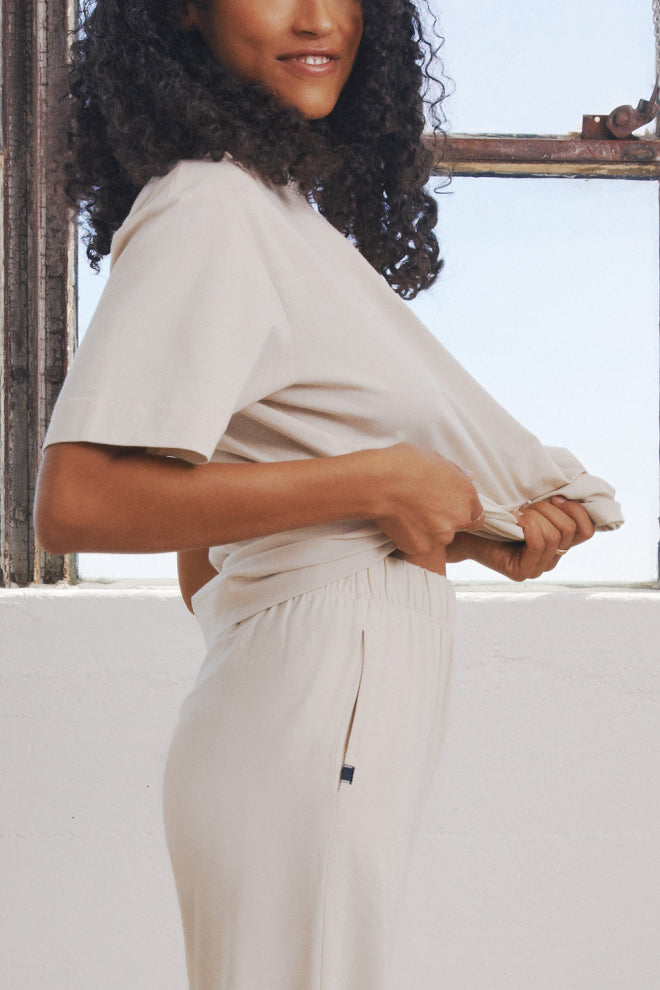 Side view of a model wearing the Perfect Pima T-Shirt and Pants in Heather Oatmeal. The model is lifting the t-shirt slightly, showcasing the soft and relaxed fit of both the top and the elastic waistband of the pants, set against a bright background with natural light streaming through a window.