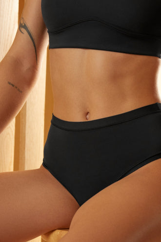 A close-up shot of a model seated against a wooden backdrop, wearing the Bliss Highwaist Brief and Bliss Bralette in Black.