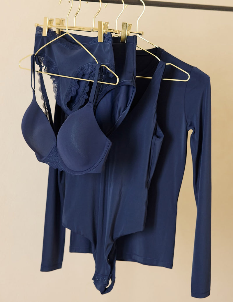 The Base T-Shirt Bra, Bliss High-Waist Brief, Bliss Bodysuit, and Bliss Long-Sleeve T-Shirt in Navy hanging on gold hangers against a neutral wall.