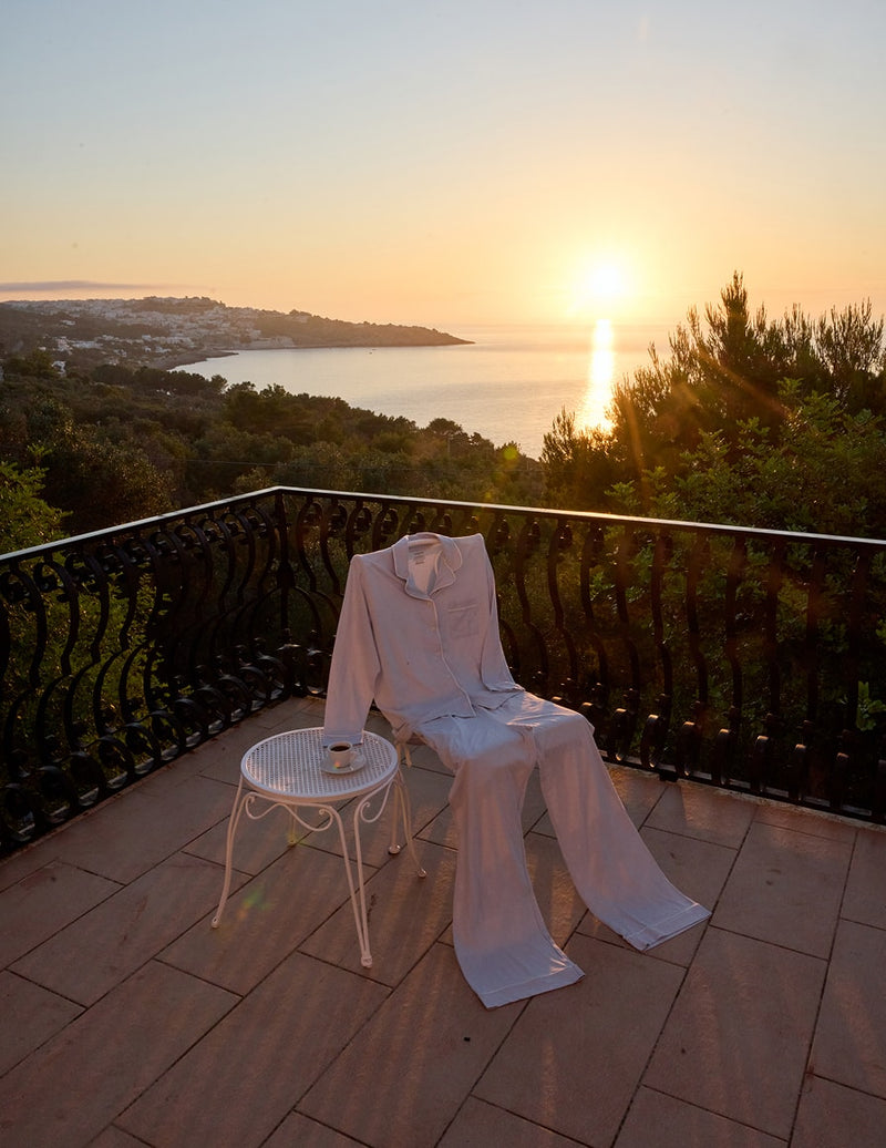 The Pima Pajama Shirt and Pants in Soft Blue draped over a chair on a terrace at sunset overlooking the Mediterranean.