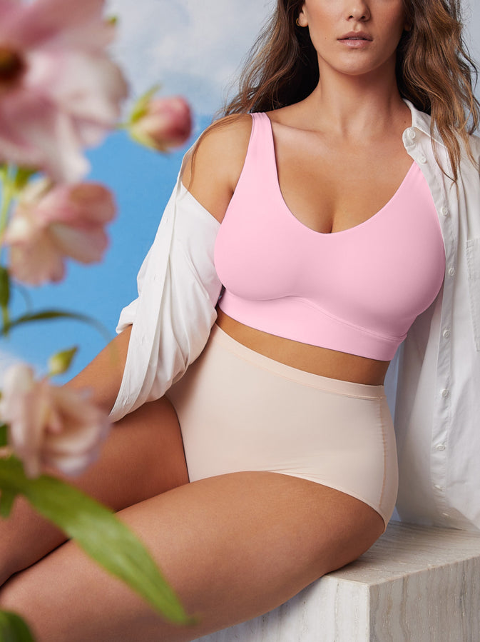 Model against a blue sky cloudy backdrop. She's wearing the Bliss Bralette in Pink with a pink flower blurry in the foreground.