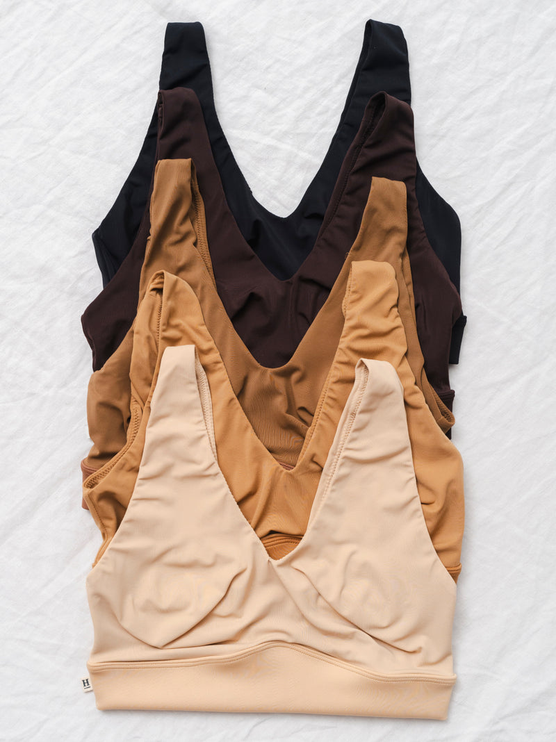 Five Bliss Bralettes are neatly arranged in a stack on a white fabric background. The bralettes are shown in the colors Black, Earth, Brown, Tan, and Beige, highlighting the variety of shades available in the collection.