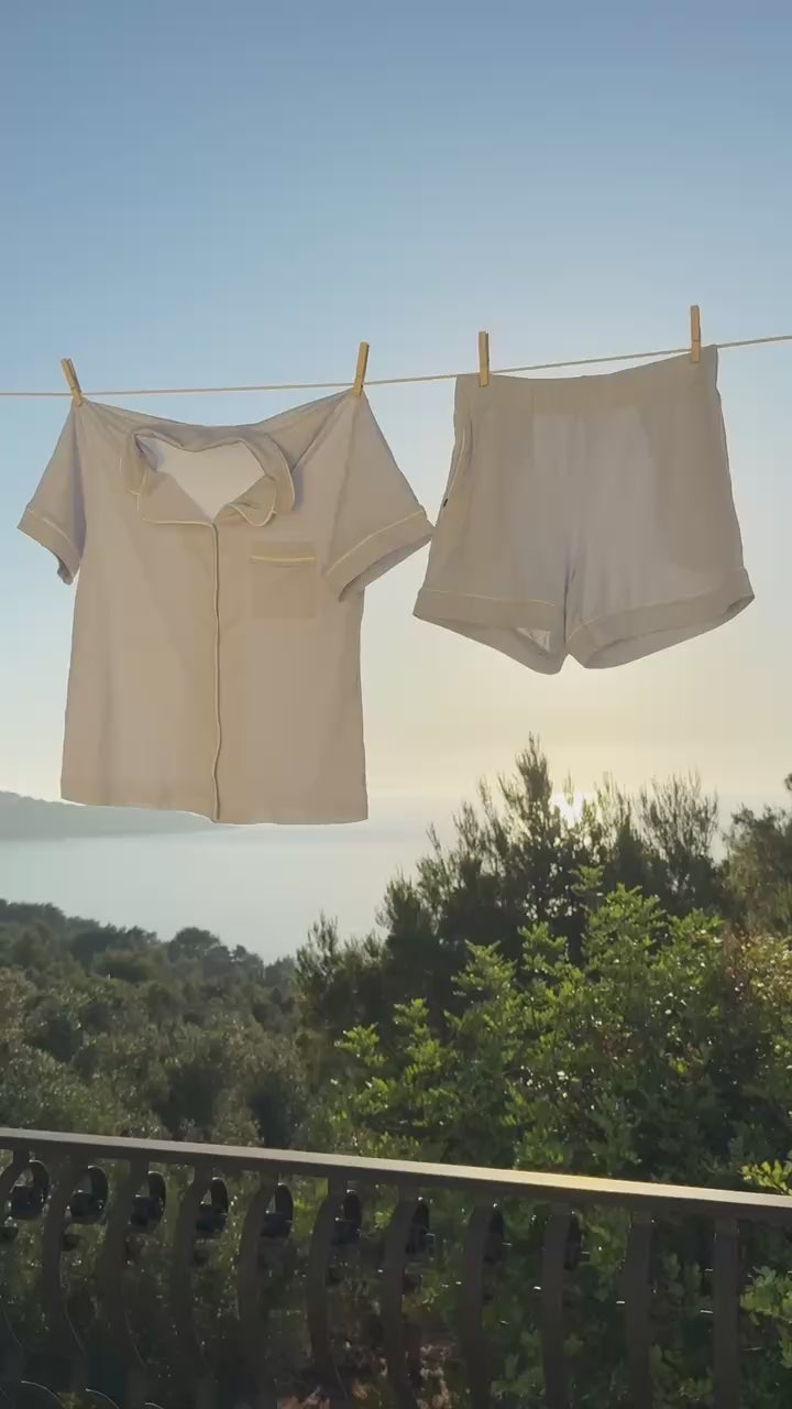 The Pima Pajama Shirt and Shorts in Soft Blue hanging on an outdoor clothesline against a coastal sunrise.