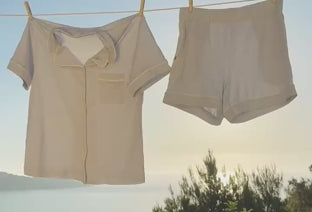 The Pima Pajama Shirt and Shorts in Soft Blue hanging on an outdoor clothesline against a coastal sunrise.